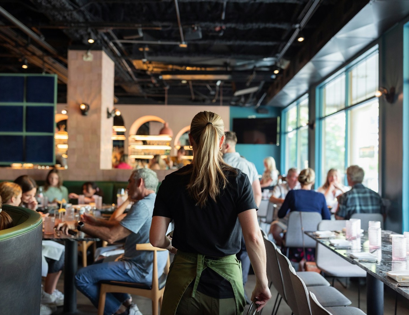 A server walking through a full restaurant dining room mid-service.